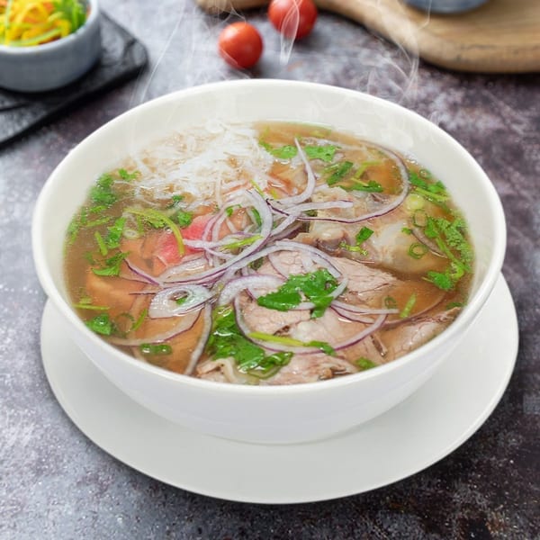 Steaming bowl of Vietnamese pho with beef, noodles, red onions, and fresh herbs.